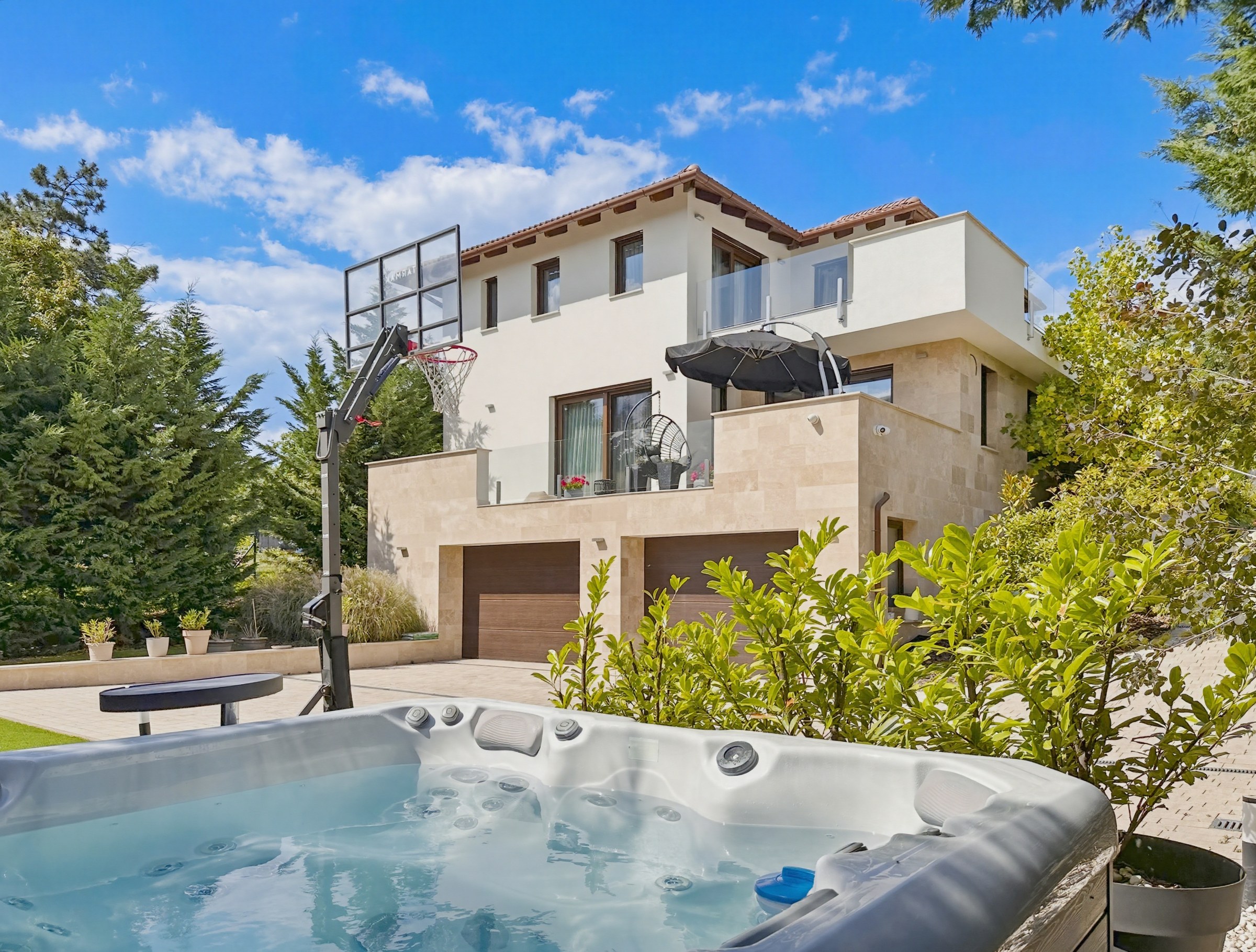 Ez a fotó egy modern, kétszintes családi házat ábrázol kívülről, gondozott kerttel, ahol egy jakuzzi és egy kosárlabda palánk is található. Az épület világos, bézs színű homlokzattal és cseréptetővel rendelkezik, több erkéllyel és garázskapukkal.
