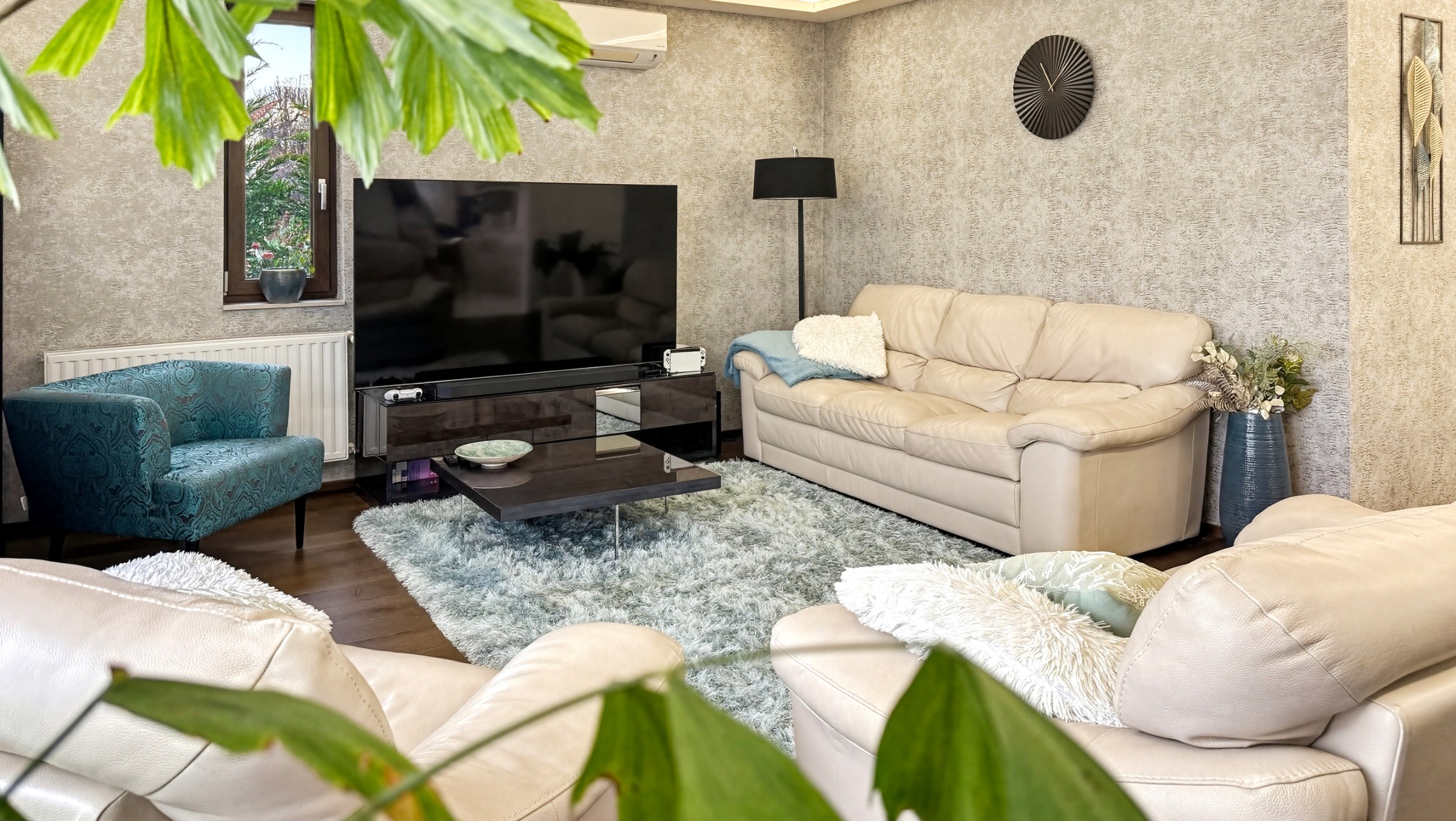Ez a nappali világos, barátságos hangulatú. Világos bőrkanapék és egy türkiz fotel uralják a teret, melyet egy nagyméretű TV és egy modern dohányzóasztal egészít ki. A falak textúrált tapétával, az ablakon át pedig természetes fény áramlik be.